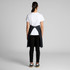 A person wearing a black Carrie Half Apron over a white T-shirt and black pants, facing away from the camera.