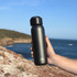 A drink bottle in matte black with a screw top lid, held against a coastal backdrop. Features branding logo.