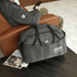 A grey duffel bag with multiple pockets and a logo, resting on a table next to a person sitting on a couch.