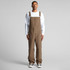 Khaki canvas overalls worn by a model, featuring adjustable straps and a relaxed fit. The model pairs them with a white long-sleeve shirt.