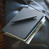 A black notebook with a pen rests on a black leather surface, featuring a textured cover and contrasting inner pages.