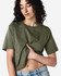 A woman in an olive green boxy T-shirt, styled with high-waisted jeans and curly hair. The shirt has a logo.