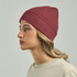 A maroon ribbed beanie worn by a model with wavy hair, facing to the side. The product has a logo.