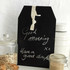 A black chalkboard sign with a tag shape, featuring handwritten messages and a hanging ribbon.