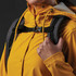 A person wearing a yellow rain jacket is holding a grey backpack strap. The backpack features a logo.