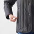 A thermal jacket in grey featuring a quilted design and a hand reaching into a side pocket. It has a logo.