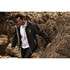 A man wearing a black softshell jacket with a logo, walking along a rocky coastline.
