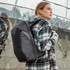 A grey backpack with a sleek design is worn by a woman in a checked coat, set against a reflective background.