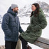 A man in a navy puffer jacket and a woman in a green puffer coat stand together outdoors in a snowy landscape.