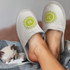 A pair of white waffle slippers with a decorative logo, resting beside a sleeping kitten on a plaid blanket.