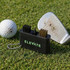 A black golf club cleaning tool with brushes sits on grass next to a golf ball and a club with dirt on it.