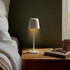 A white rechargeable table lamp stands on a wooden bedside table, softly glowing next to a pair of glasses and a book.