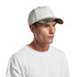 A white cap with a camo-patterned peak, featuring a logo on the front, worn by a model.