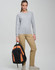 A young woman stands with an orange and black backpack, wearing a grey long-sleeve top and beige trousers.