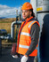 A man wearing an orange hi-vis hooded puffer vest stands beside a metallic structure, holding a laptop.