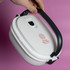 A white lunch box with a black handle, featuring a floral logo, held against a pink background.