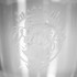 A glass cup with an etched design featuring a campervan, mountains, and waves, along with a logo.