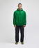 A green pullover hooded sweatshirt with a front pocket, paired with black trousers. The model stands against a neutral background.