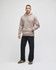 A man stands wearing a beige hooded sweatshirt and dark jeans, with sneakers. The sweatshirt has a logo.