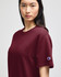A maroon short-sleeve t-shirt featuring a logo on the sleeve. The model presents the shirt in a neutral setting.