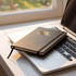 A5 weekly diary in black with gold initials, resting on a laptop keyboard by a sunlit window.