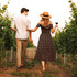 A couple walks through a vineyard, the woman in a floral dress holding a wine glass and wicker basket. Sunlight filters through the vines.