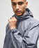 A grey full-zip anorak jacket featuring a hood and a logo on the sleeve, modelled by a young man.