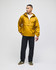 A man wearing a yellow packable anorak jacket stands with black pants and green shoes. The jacket has a logo.