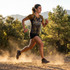 A woman runs on a dirt path, wearing a yellow and black active V-neck t-shirt and shorts, surrounded by trees and mountains.