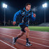 A man in a black and blue active hoodie runs on a track under stadium lights, wearing shorts and athletic socks.