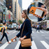 A woman in a navy suit walks across a city street, holding an orange MagSafe power bank and smartphone.