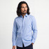 A man in a light blue long-sleeve shirt stands against a neutral background. The shirt features a collar and buttons.