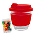 A reusable glass coffee cup with a red silicone lid and sleeve, alongside a packet of colourful jelly beans.