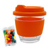 A glass coffee cup with an orange silicone lid and sleeve, accompanied by a pack of colourful jelly beans.