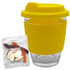 A reusable glass coffee cup with a yellow silicone lid and sleeve, accompanied by a clear bag of assorted jelly sweets.