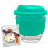 A clear glass coffee cup with a teal silicone lid and sleeve, accompanied by a packet of mixed lollies.