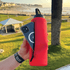 A red drink bottle with a zipped compartment, holding a smartphone, set against a scenic beach background.