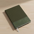 A black A5 notebook with a textured cover, resting on a wooden desk beside a window.