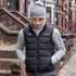 A black unisex puffer vest worn over a grey sweater, paired with a grey beanie. The vest features a logo.