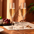 Two elegant, clear champagne flutes filled with sparkling wine, positioned on a wooden table with grapes and nuts.