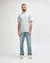A man stands wearing a light grey Gildan Softstyle adult midweight T-shirt and blue jeans. The shirt has a logo.