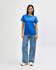 A woman stands wearing a blue Gildan Softstyle ladies midweight T-shirt and jeans, paired with white sneakers.