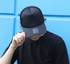 A black trucker cap with a mesh back, featuring a logo, worn by a person against a blue background.