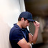 A young man in a navy cap leans against a wall, resting his hand on the brim, appearing thoughtful.
