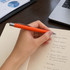 An orange metal pen held above a notepad with handwritten meeting notes, placed on a wooden desk near a laptop.