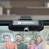 A black Helm Webcam Camera with a logo, attached to a monitor, captures a family video call in the background.