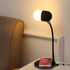 A black and white wireless charging sound lamp with a flexible neck, next to a smartphone on a charging pad.