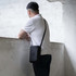 A black crossbody bag is slung over a man's shoulder while he leans against a wall.