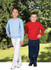Two children stand outdoors, one wearing a light blue jumper and the other in a bright red jumper, both smiling.