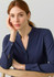 A woman poses wearing a navy long sleeve blouse with a button-up neckline, sitting against a light background.